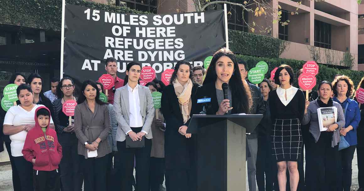 A woman with a microphone stands in front of a group of roughly twenty people and a sign that reads "15 MILES SOUTH OF HERE REFUGEES ARE DYING AT THE BORDER"  