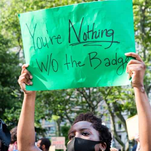 A masked person raises a green posterboard wtih the words "You're Nothing W/O the Badge"